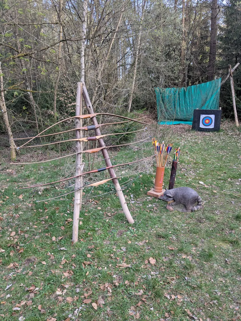 Selbstgebaute Bögen lehnen an einem Gestell aus Ästen, dahinter eine Zielscheibe